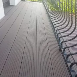 Nowoczesny taras z desek kompozytowych w odcieniach brązu, widok z góry na balustradę z metalowymi zdobieniami, jasne tło ściany budynku.