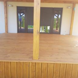 DachNel - Nowa drewniana podłoga tarasu z deskami w ciepłym odcieniu brązu, z drewnianymi słupami podtrzymującymi zadaszenie, widok z Ostrzeszowa na okna z roletami zewnętrznymi.