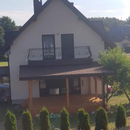 DachNel - Dom jednorodzinny z nowym, brązowym dachem z dachówki bitumicznej, balkonem z ozdobną balustradą i drewnianą werandą, otoczony zielenią.