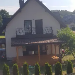 Dom jednorodzinny z nowym, brązowym dachem z dachówki bitumicznej, balkonem z ozdobną balustradą i drewnianą werandą, otoczony zielenią.