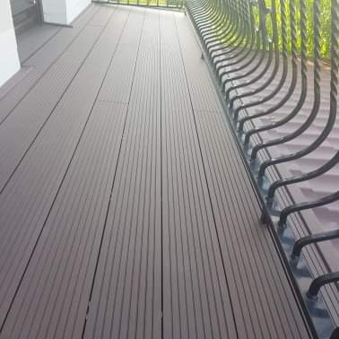 Nowoczesny taras z desek kompozytowych w odcieniach brązu, widok z góry na balustradę z metalowymi zdobieniami, jasne tło ściany budynku.