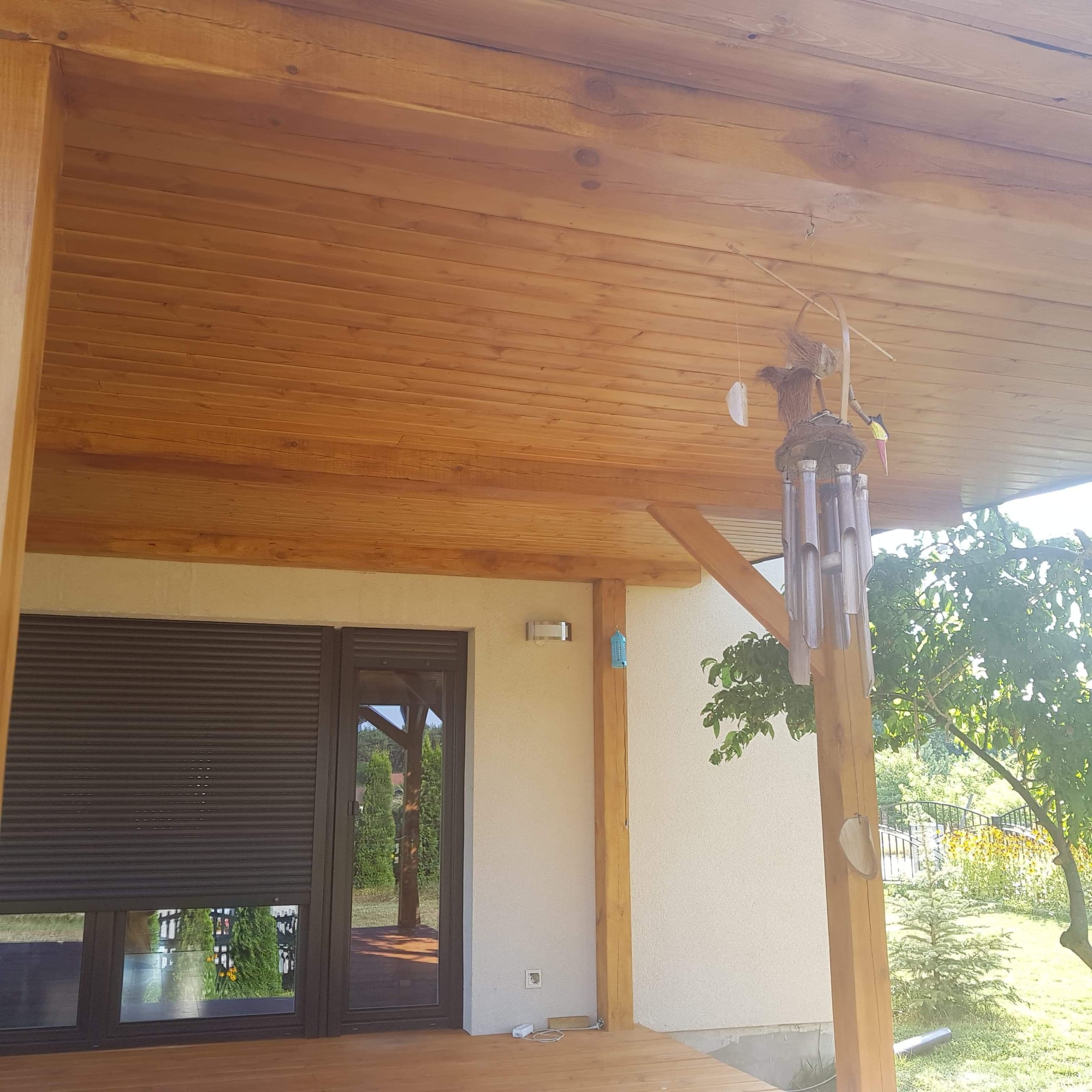 Drewniany taras z zadaszeniem z desek, widoczne okno z roletami zewnętrznymi, drewniane słupy i dekoracja wisząca na słupie, w tle zieleń ogrodu.