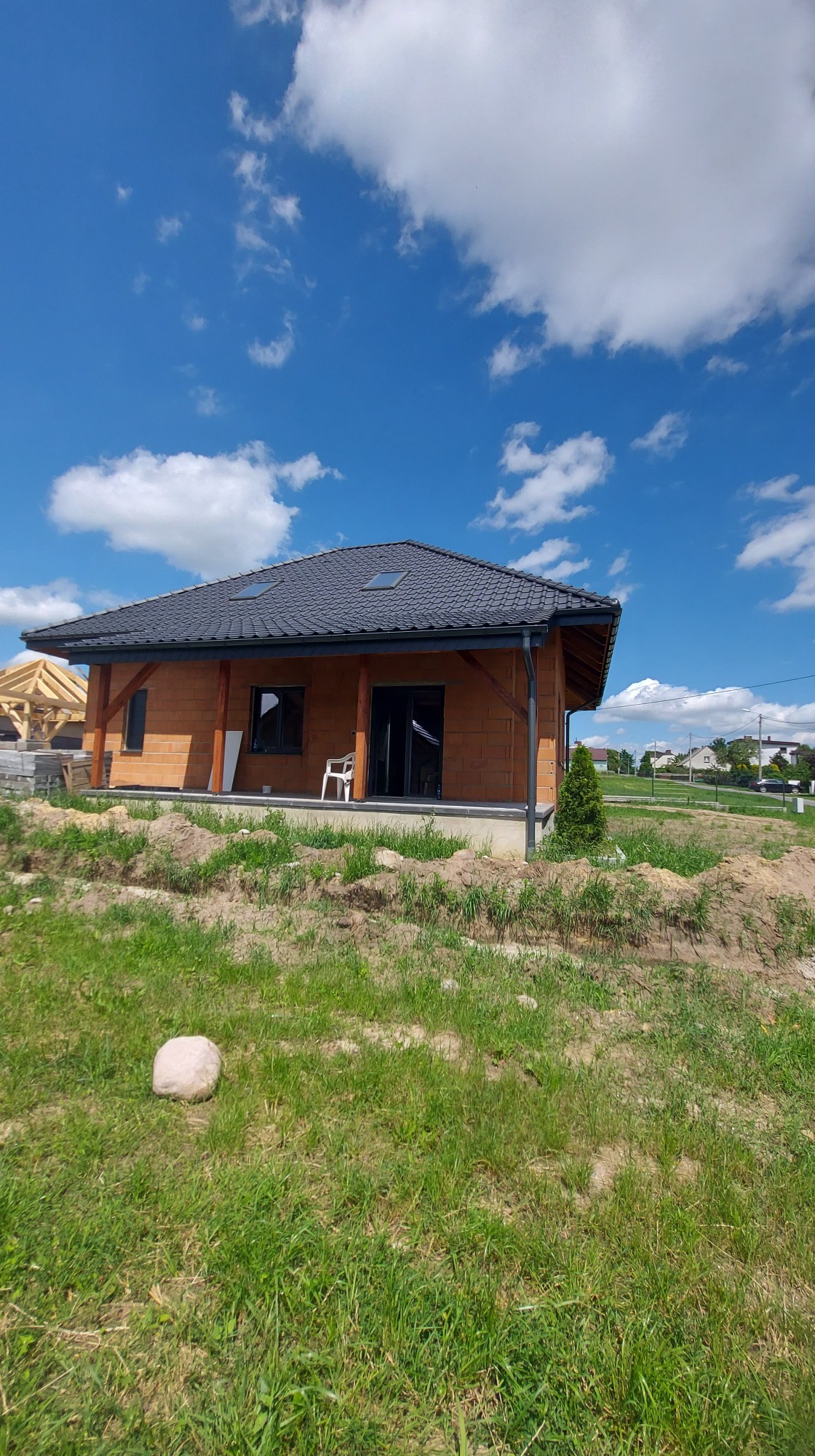 Nowoczesny dom jednorodzinny z ciemną dachówką, widoczne okna dachowe, drewniane elementy konstrukcyjne i częściowo zagospodarowany teren wokół.