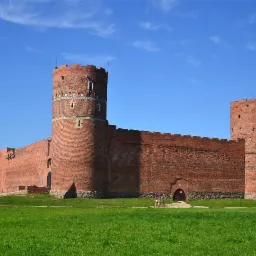 Średniowieczny zamek z czerwonej cegły z dwiema okrągłymi wieżami i fragmentem muru obronnego, otoczony zielonym trawnikiem pod błękitnym niebem.