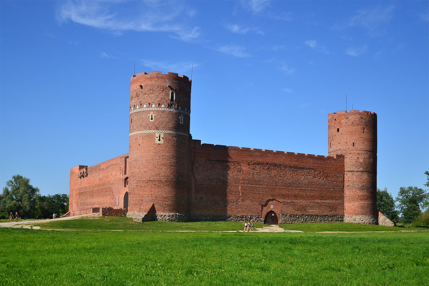 Średniowieczny zamek z czerwonej cegły z dwiema okrągłymi wieżami i fragmentem muru obronnego, otoczony zielonym trawnikiem pod błękitnym niebem.