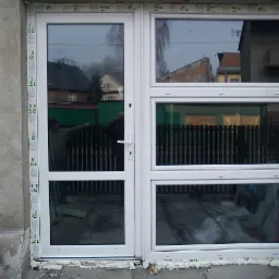 Białe okno PCV z drzwiami balkonowymi, zamontowane w surowym otworze ściany, widoczne ślady pianki montażowej i wiadro z masą szpachlową obok.