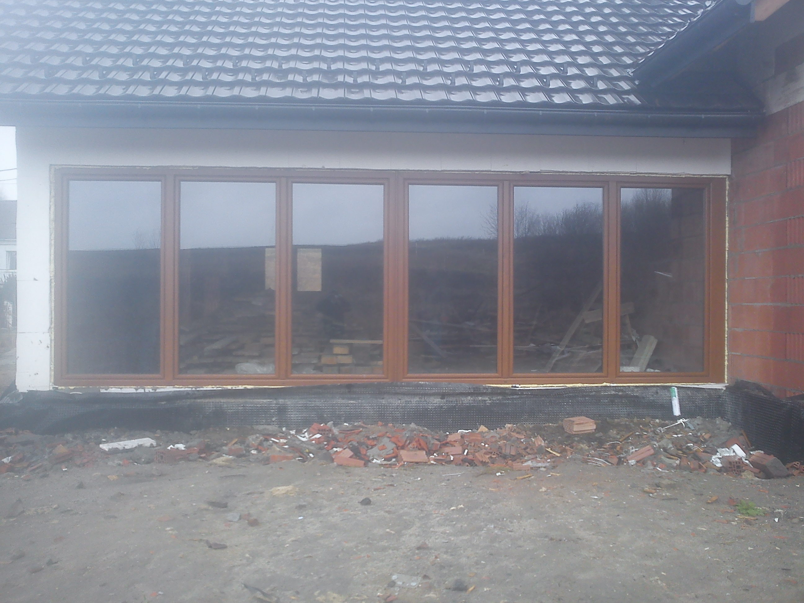 Montaż dużych, brązowych okien w surowym stanie budynku, widoczne ślady prac murarskich i gruz przed oknami.