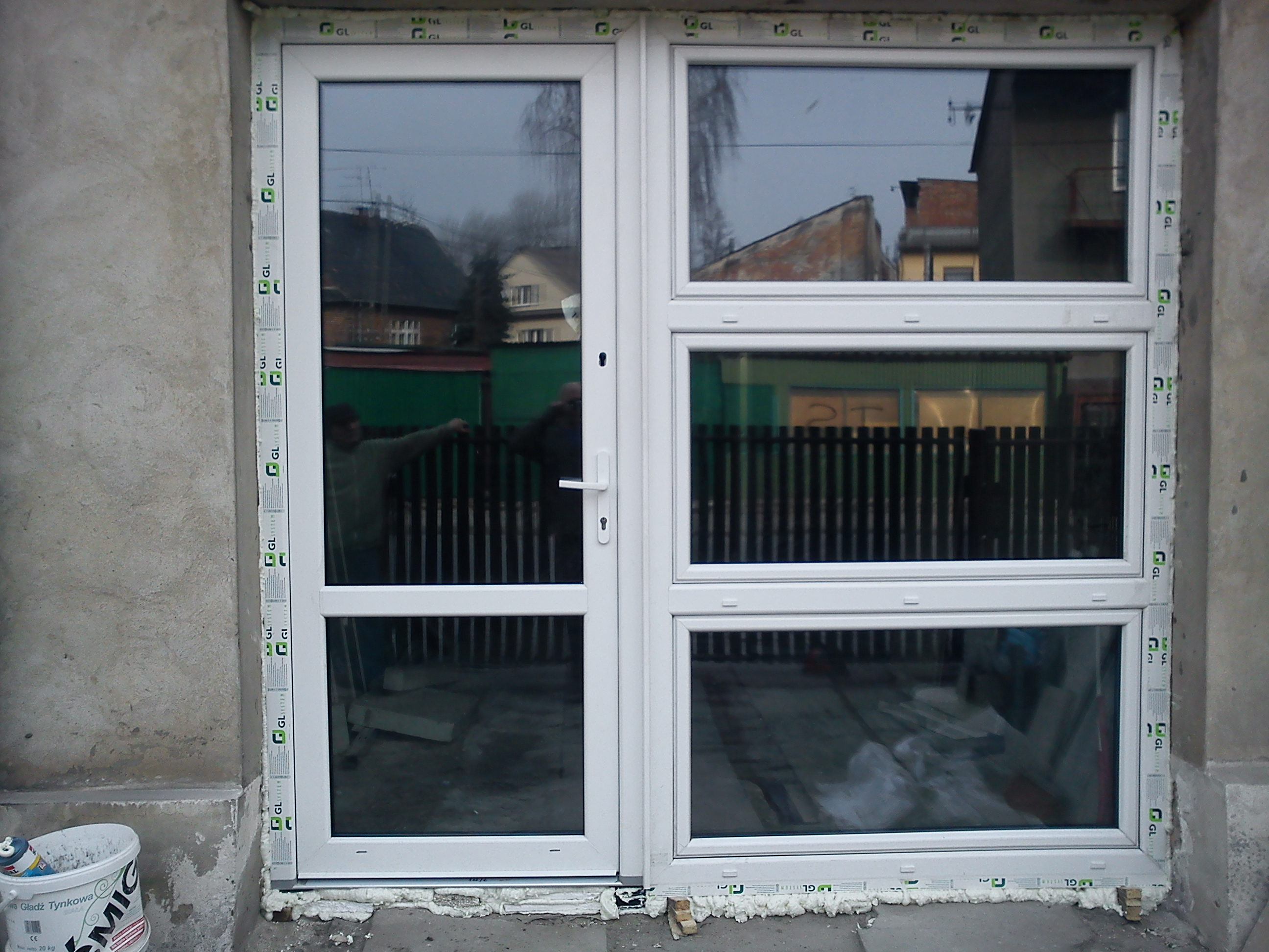 Białe okno PCV z drzwiami balkonowymi, zamontowane w surowym otworze ściany, widoczne ślady pianki montażowej i wiadro z masą szpachlową obok.