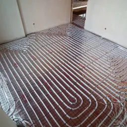 Białe rurki systemu rozprowadzenia ciepła ułożone w pętle na metalowej siatce, instalacja w surowym stanie, widoczne przyłącza niebieskie i czerwone.