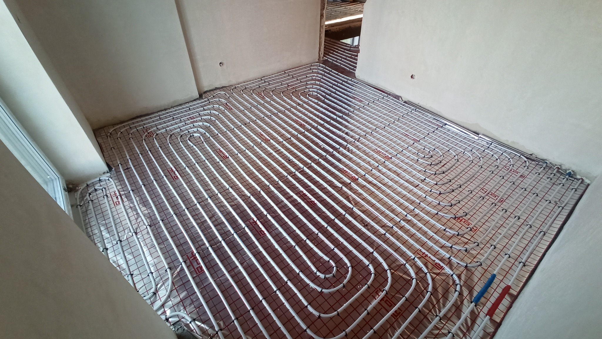 Białe rurki systemu rozprowadzenia ciepła ułożone w pętle na metalowej siatce, instalacja w surowym stanie, widoczne przyłącza niebieskie i czerwone.