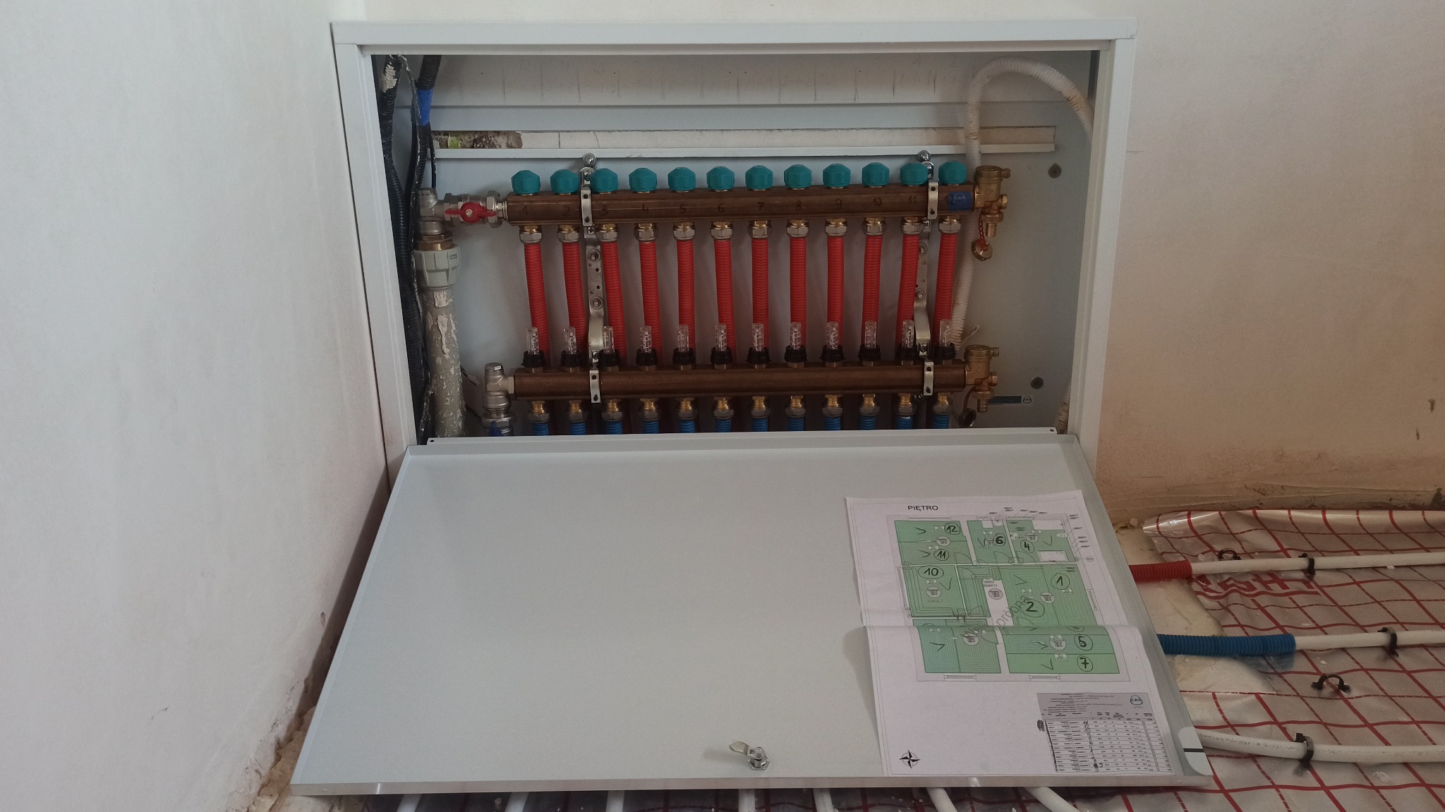 Rozdzielacz ogrzewania podłogowego Kan-Therm w szafce z otwartymi drzwiczkami, widoczny schemat instalacji i rury w izolacji na podłodze.