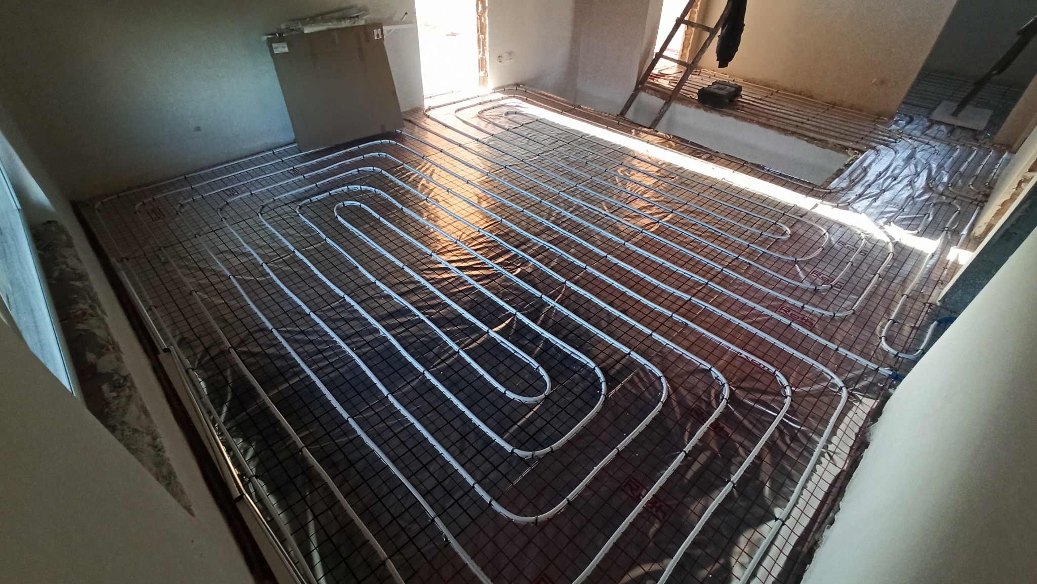 Białe rurki systemu rozprowadzenia ciepła ułożone na czarnej folii z siatką, przygotowane do zalania betonem w nowym budynku.