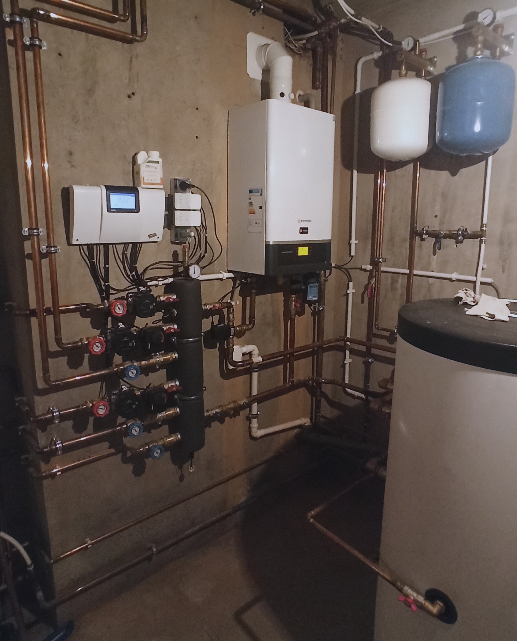 Kondensacyjny kocioł gazowy Warmhaus Enerwa Plus z trzema obiegami grzewczymi, sterowanymi pogodowo, zamontowany na ścianie w kotłowni, widoczne liczne miedziane rury, zawory i manometry.