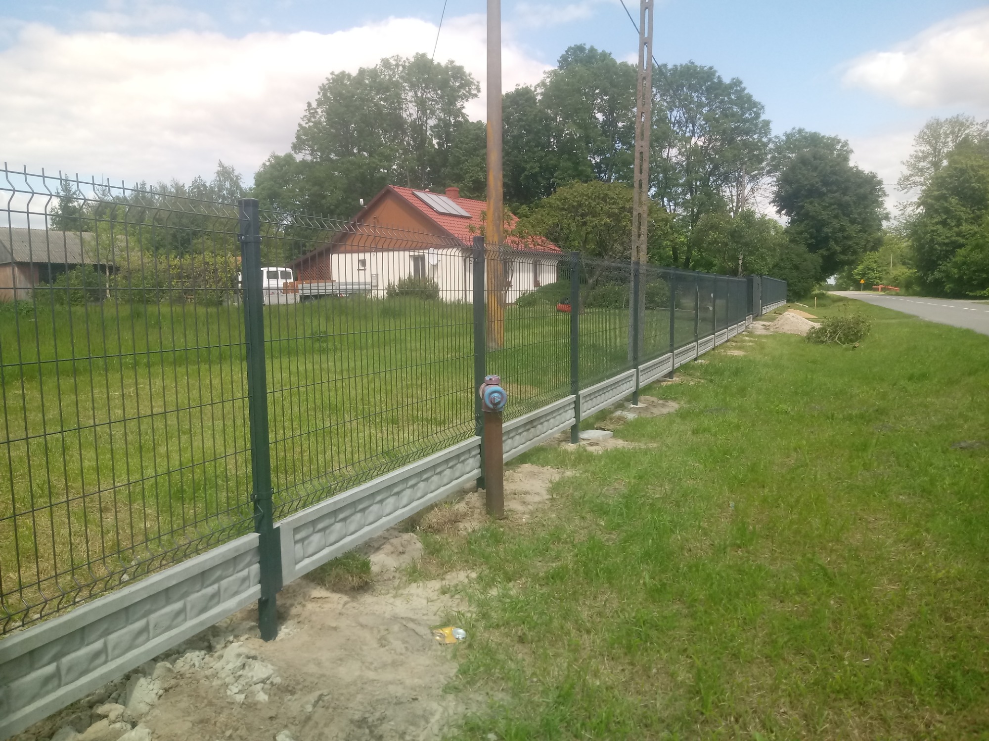 Nowo postawione ogrodzenie panelowe z podmurówką imitującą cegłę, widoczne na tle domu z czerwonym dachem i zieleni, z elementami wykończeniowymi w trakcie montażu.