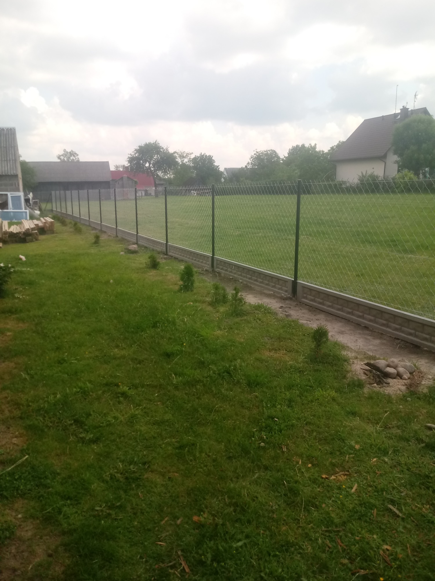 Długi, zielony panelowy płot z podmurówką betonową, oddzielający posesję z domami od rozległego trawnika, widziany z perspektywy trawy. W tle sterta drewna.