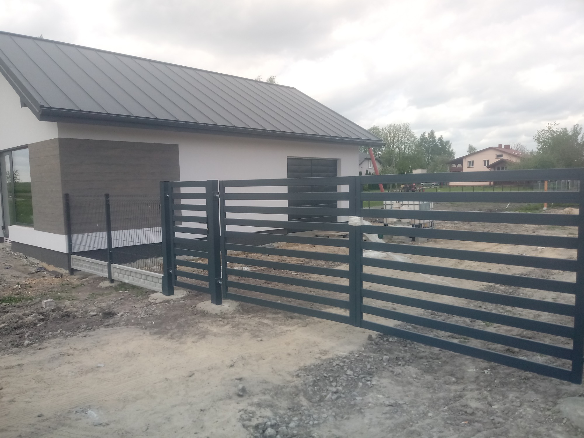Nowoczesne ogrodzenie posesji z poziomych, szarych profili stalowych, z bramą wjazdową, na tle domu z szarą blachodachówką i fragmentem ogrodzenia panelowego.