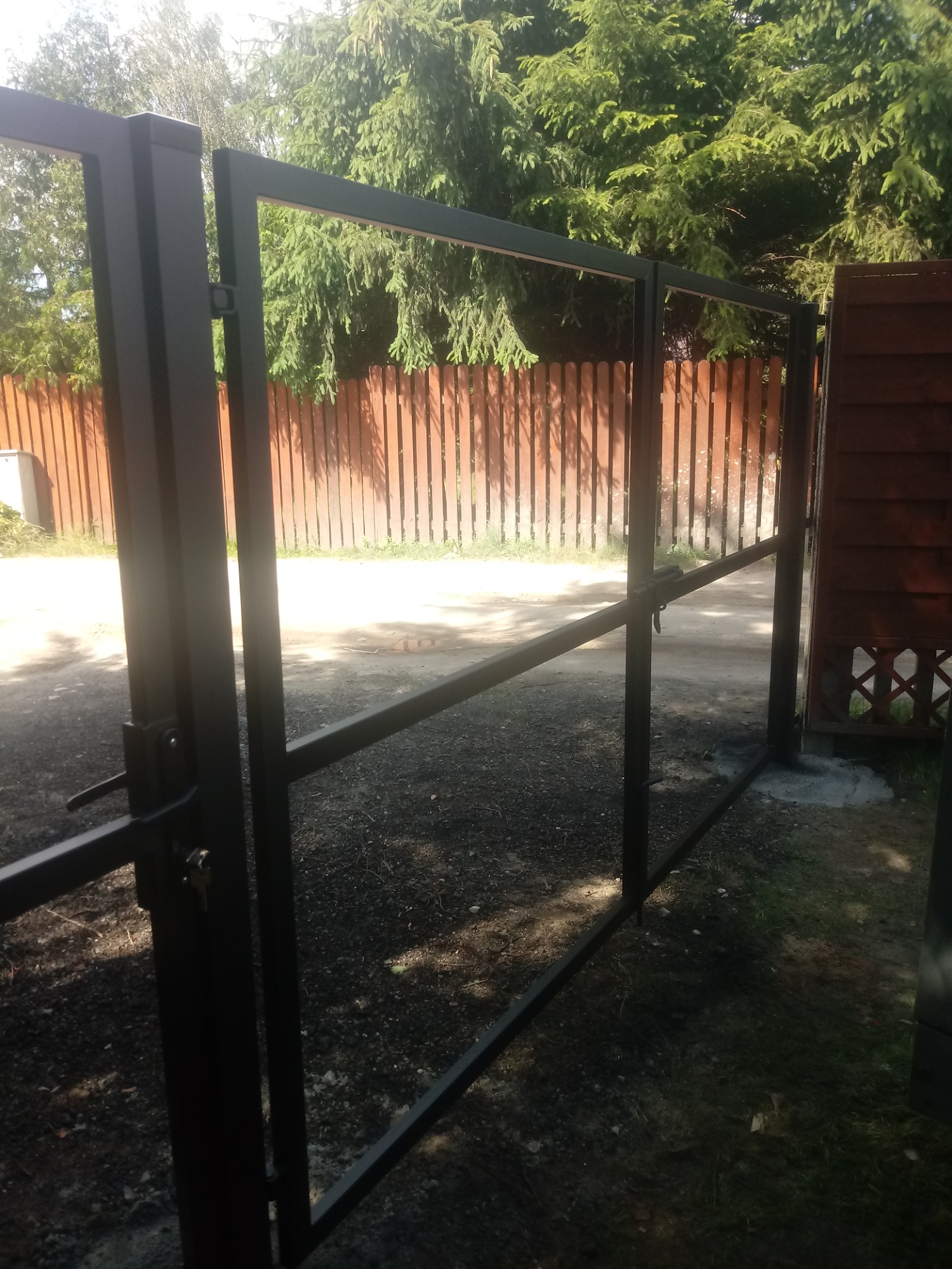 Otwarta, czarna, metalowa brama wjazdowa z widocznym zamkiem i kluczem, na tle drewnianego ogrodzenia i zieleni.