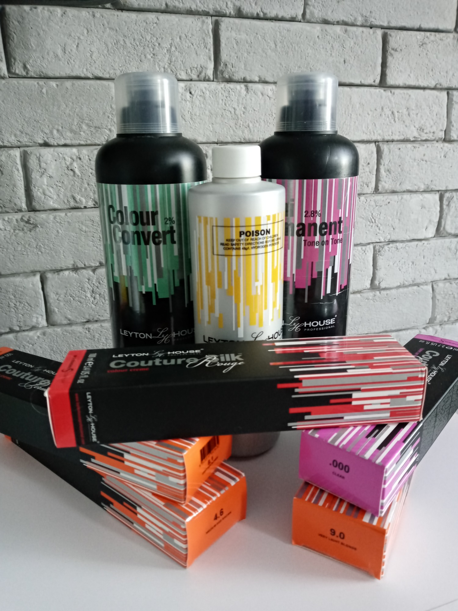 Zestaw kosmetyków do koloryzacji włosów marki Leyton House: farby w kartonikach z numeracją kolorów i butelki z preparatami Colour Convert i Permanent Tone on Tone na tle ściany z białej cegły.
