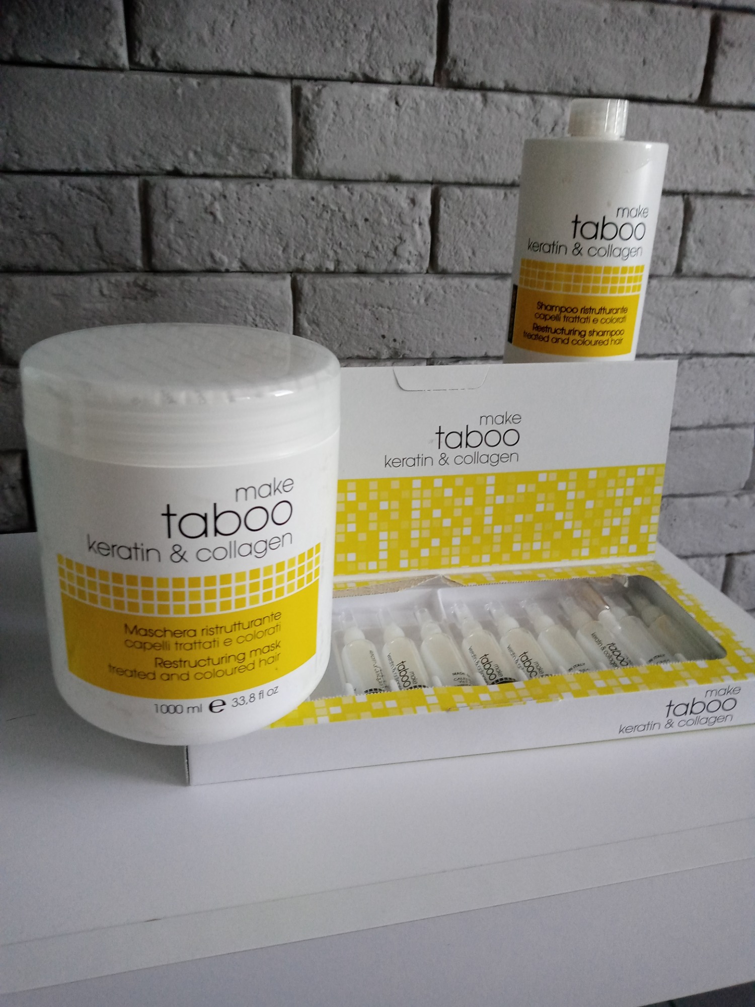 Zestaw kosmetyków do włosów marki Make Taboo: maska restrukturyzująca, szampon i ampułki z keratyną i kolagenem, na tle szarej ściany z cegieł.