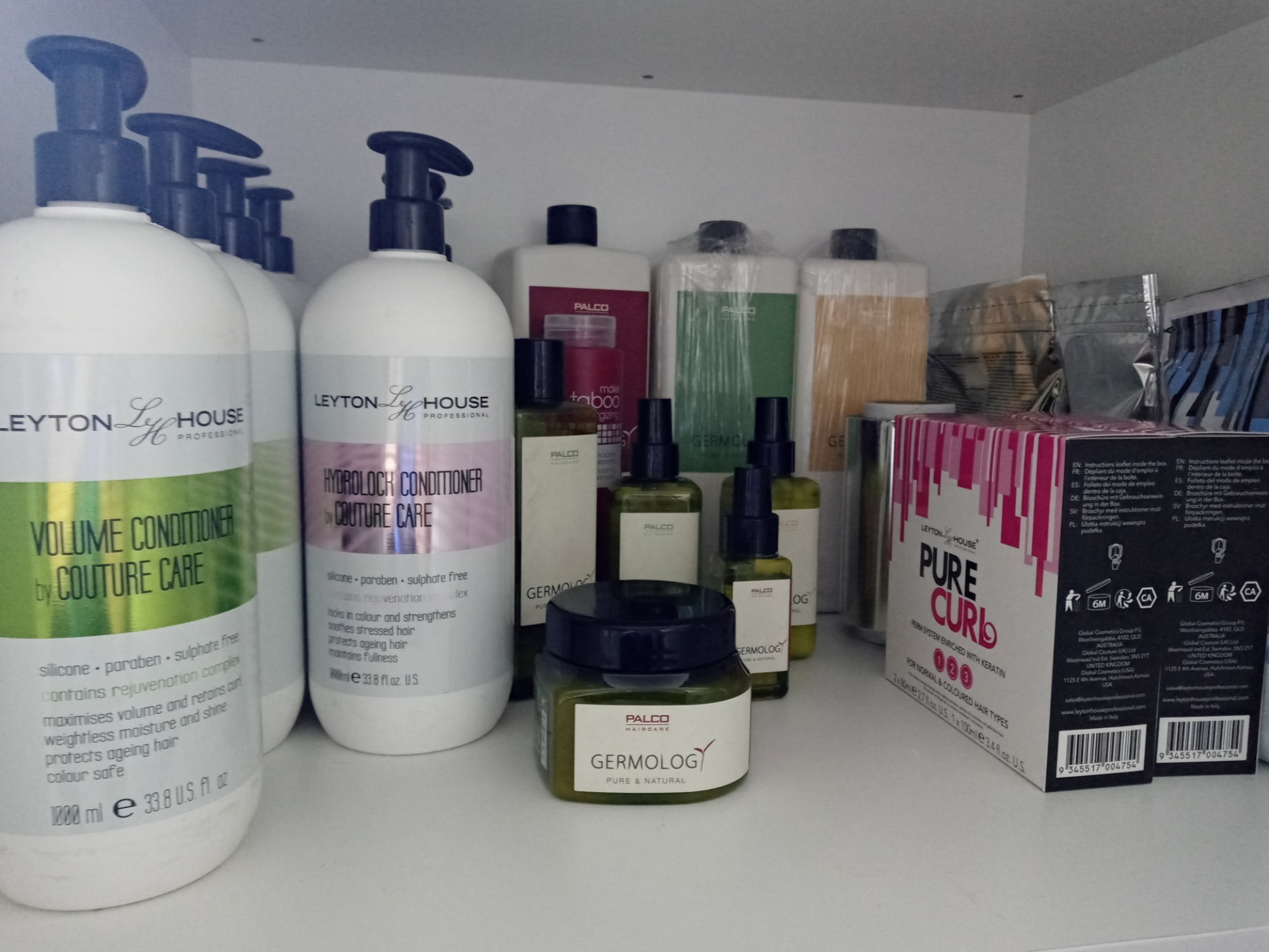 Półka z różnorodnymi produktami do pielęgnacji włosów, w tym butelki z odżywkami Leyton House, kosmetyki Palco Germology oraz zestaw do trwałej ondulacji Leyton House Pure Curl.