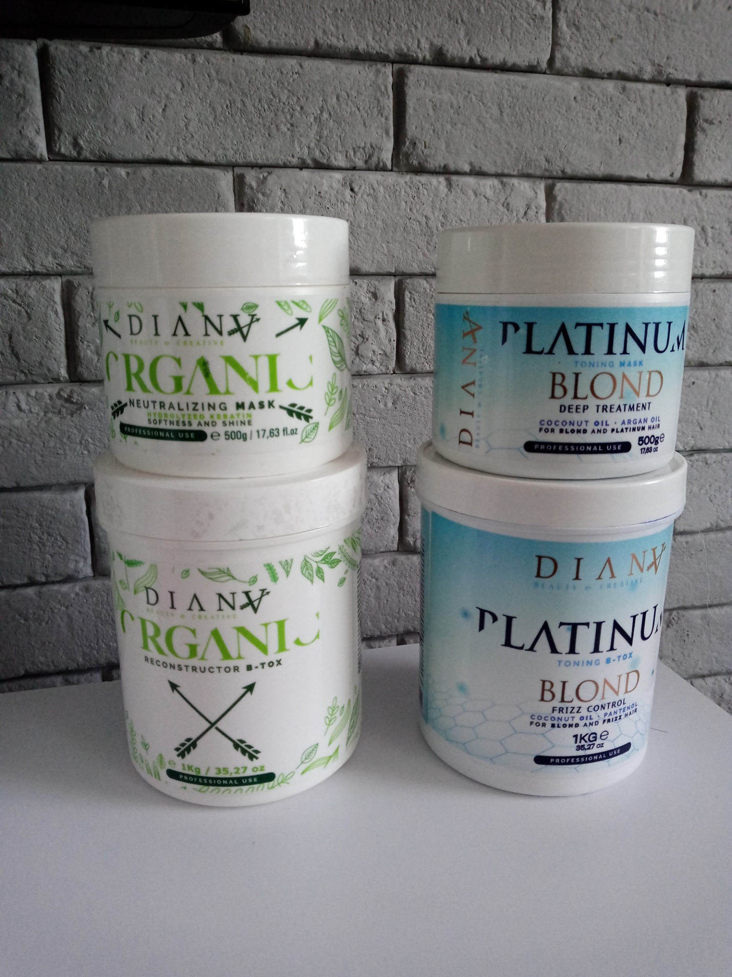 Cztery białe pojemniki z maskami do włosów marki Dianv Organics i Dianv Platinum Blond na tle szarej ściany z cegieł, widoczne opisy Neutralizing Mask, Reconstructor B-Tox i Toning Mask.