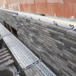 Wznoszenie ściany z cegły klinkierowej w trakcie budowy domu, widoczne rusztowanie i materiały budowlane, izolacja termiczna ściany.