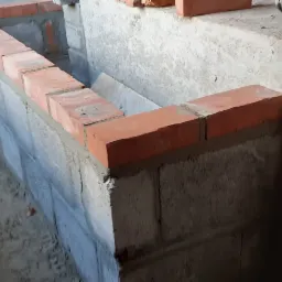 Wznoszenie ścian fundamentowych z bloczków betonowych i cegły klinkierowej, widoczne spoiny i zaprawa murarska.