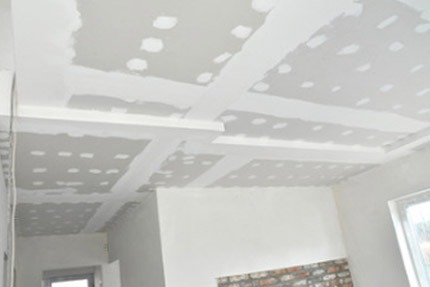 Sufit w trakcie wykańczania z płyt kartonowo-gipsowych, widoczne łączenia i miejsca po wkrętach wymagające szpachlowania, częściowo pomalowana ściana.