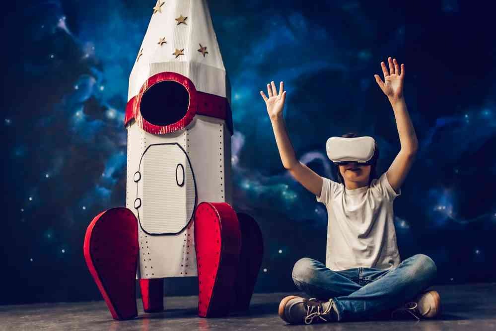 Chłopiec w okularach VR siedzi skrzyżnie obok kartonowej rakiety na tle kosmosu, ręce uniesione w górę.