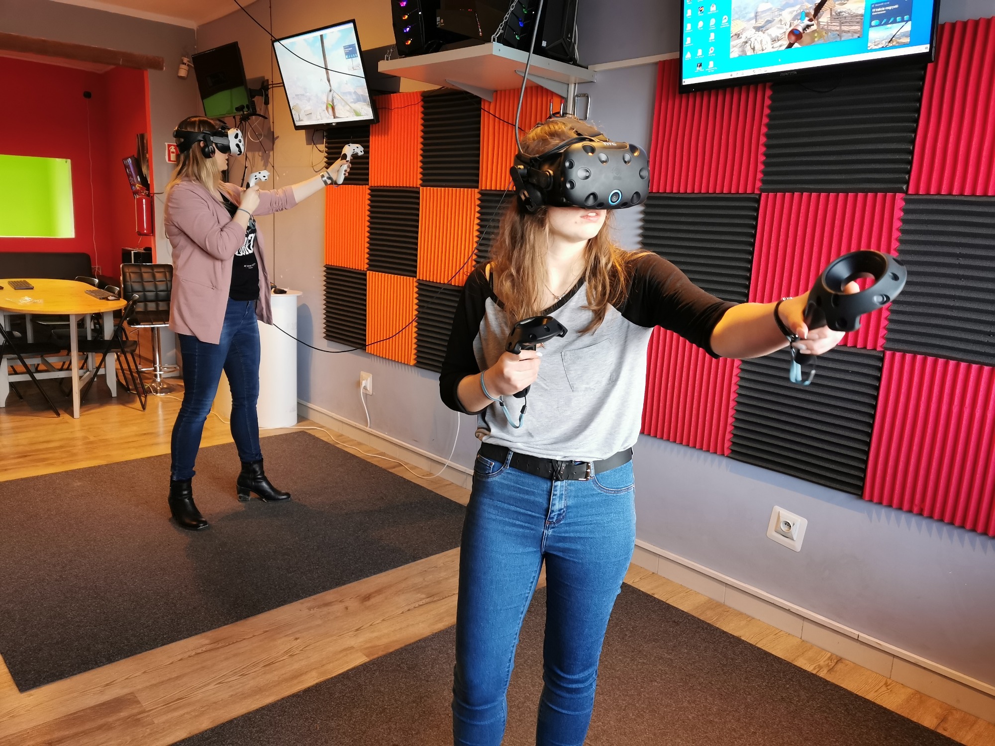 Dwie osoby w Gdyni korzystają z zestawów VR, trzymając kontrolery w dłoniach; w tle ściana wyłożona panelami akustycznymi w kolorach czerwonym i pomarańczowym.