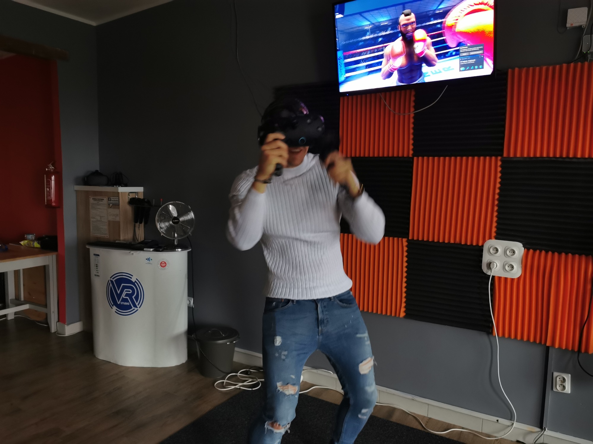 Mężczyzna w białym swetrze i podartych jeansach, z goglami VR na głowie, boksuje w wirtualnej rzeczywistości w pomieszczeniu z akustycznymi panelami i ekranem telewizora wyświetlającym grę bokserską.