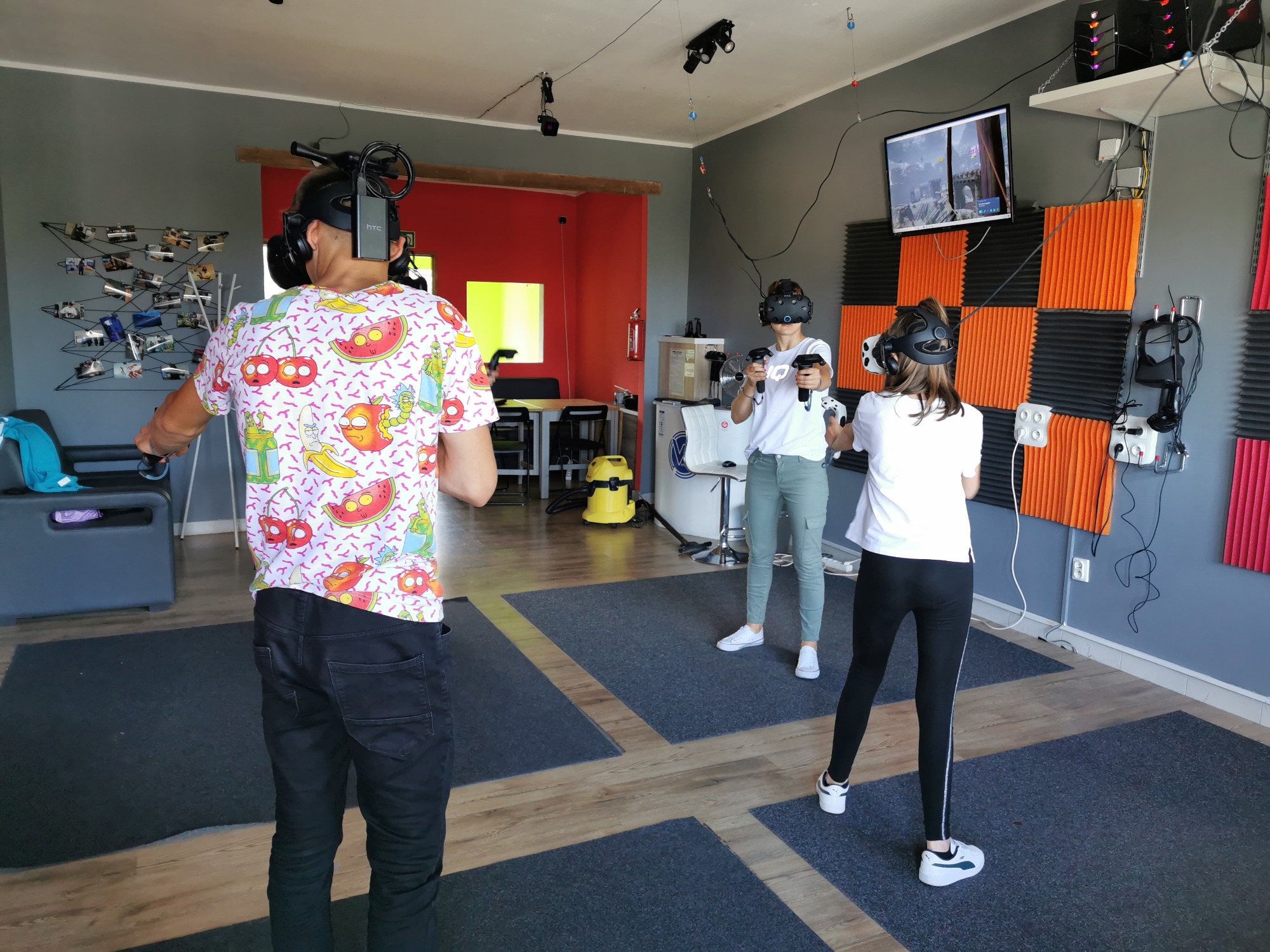 Trzy osoby w okularach VR i kontrolerach grają w grę w pomieszczeniu z szarymi ścianami, pomarańczowymi panelami akustycznymi i ekranem wyświetlającym rozgrywkę.