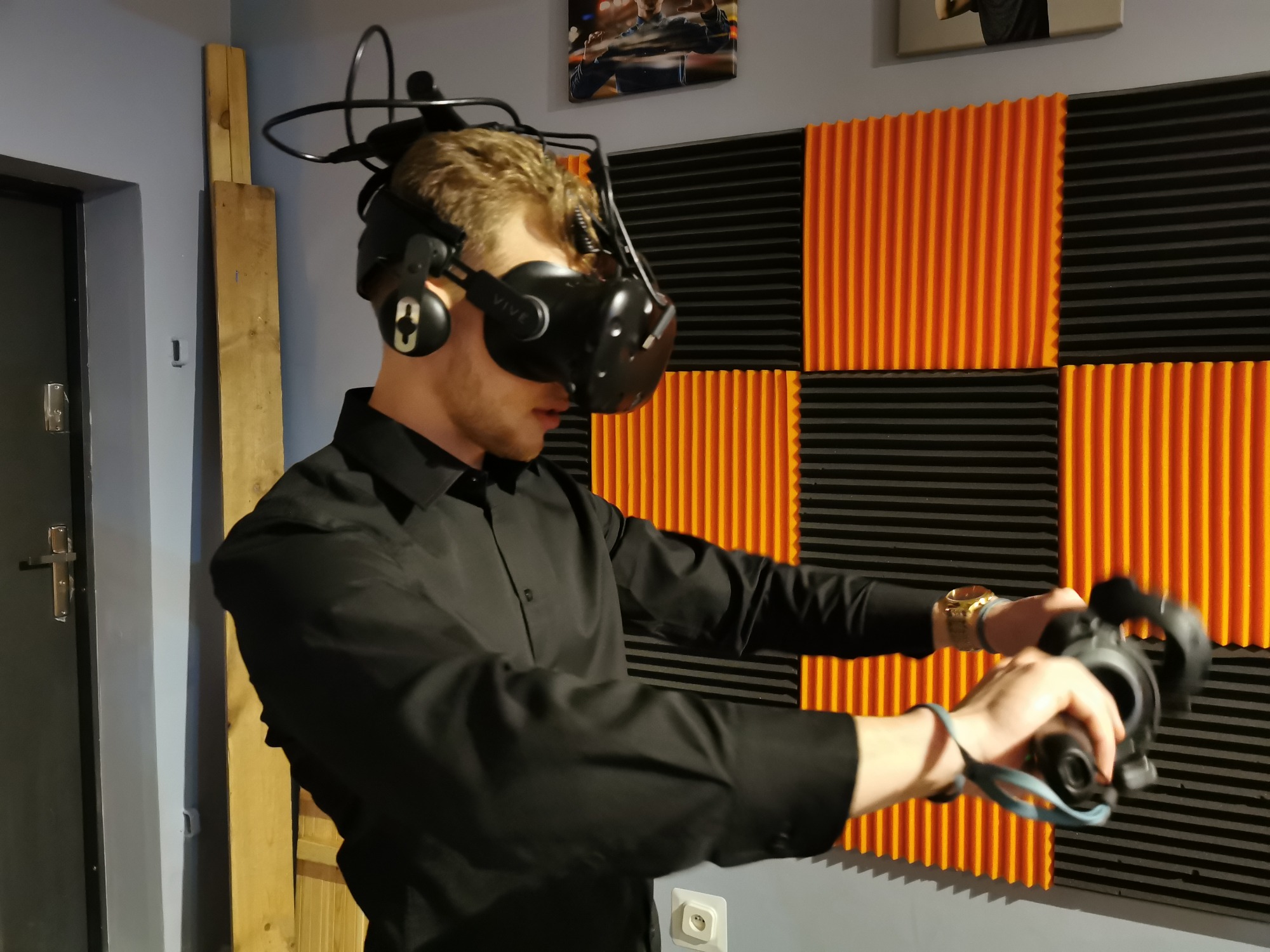 Młody mężczyzna w czarnej koszuli, używający zestawu VR Vive z kontrolerami na tle ściany z panelami akustycznymi w kolorze pomarańczowo-czarnym.