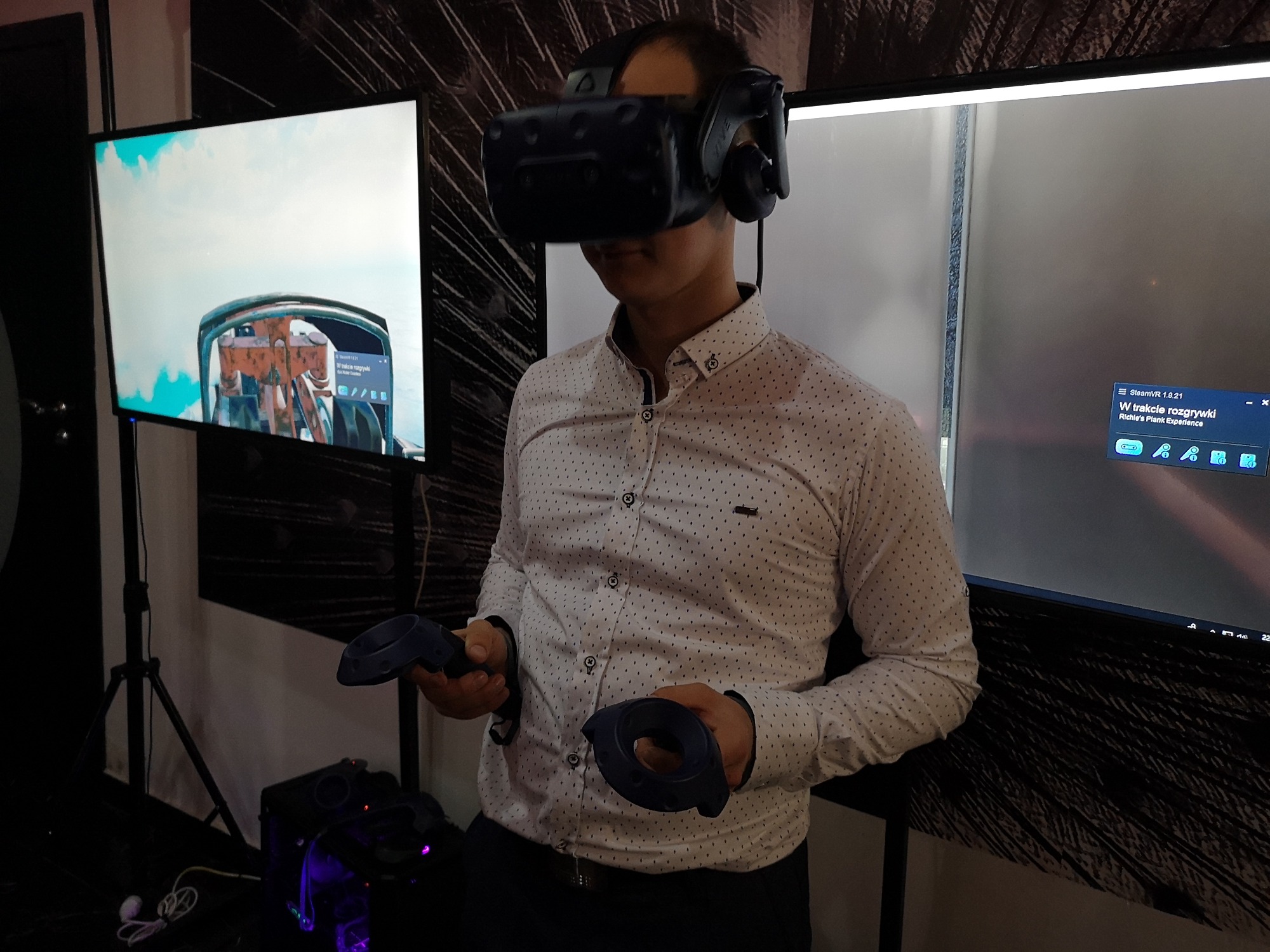 Mężczyzna w koszuli w kropki używa zestawu VR z kontrolerami, w tle ekrany wyświetlające symulację lotu i interfejs SteamVR.