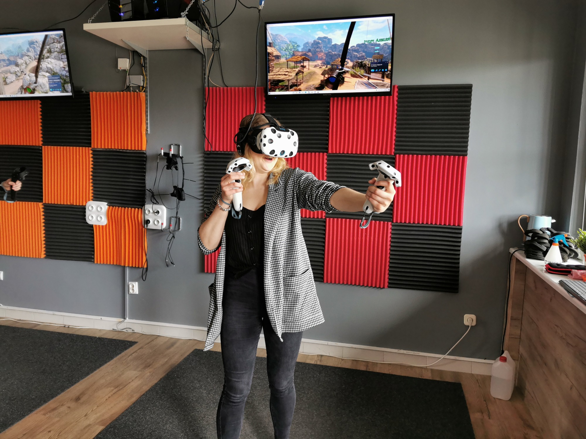 Uśmiechnięta kobieta w okularach VR trzyma kontrolery w pokoju z panelami akustycznymi i ekranem wyświetlającym grę.