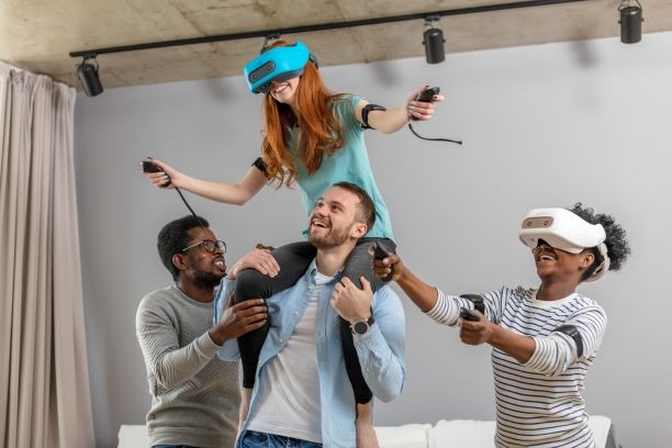 Grupa czterech uśmiechniętych osób, dwie w okularach VR, grających w grę, jedna siedzi na barana, wszyscy trzymają kontrolery.