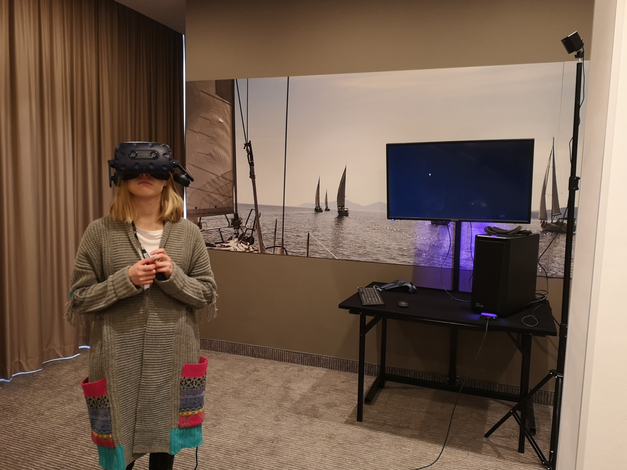 Kobieta z blond włosami w szarym swetrze z kolorowymi kieszeniami używa zestawu VR na tle ściany z motywem żaglówek, obok czarnego biurka z monitorem i komputerem.