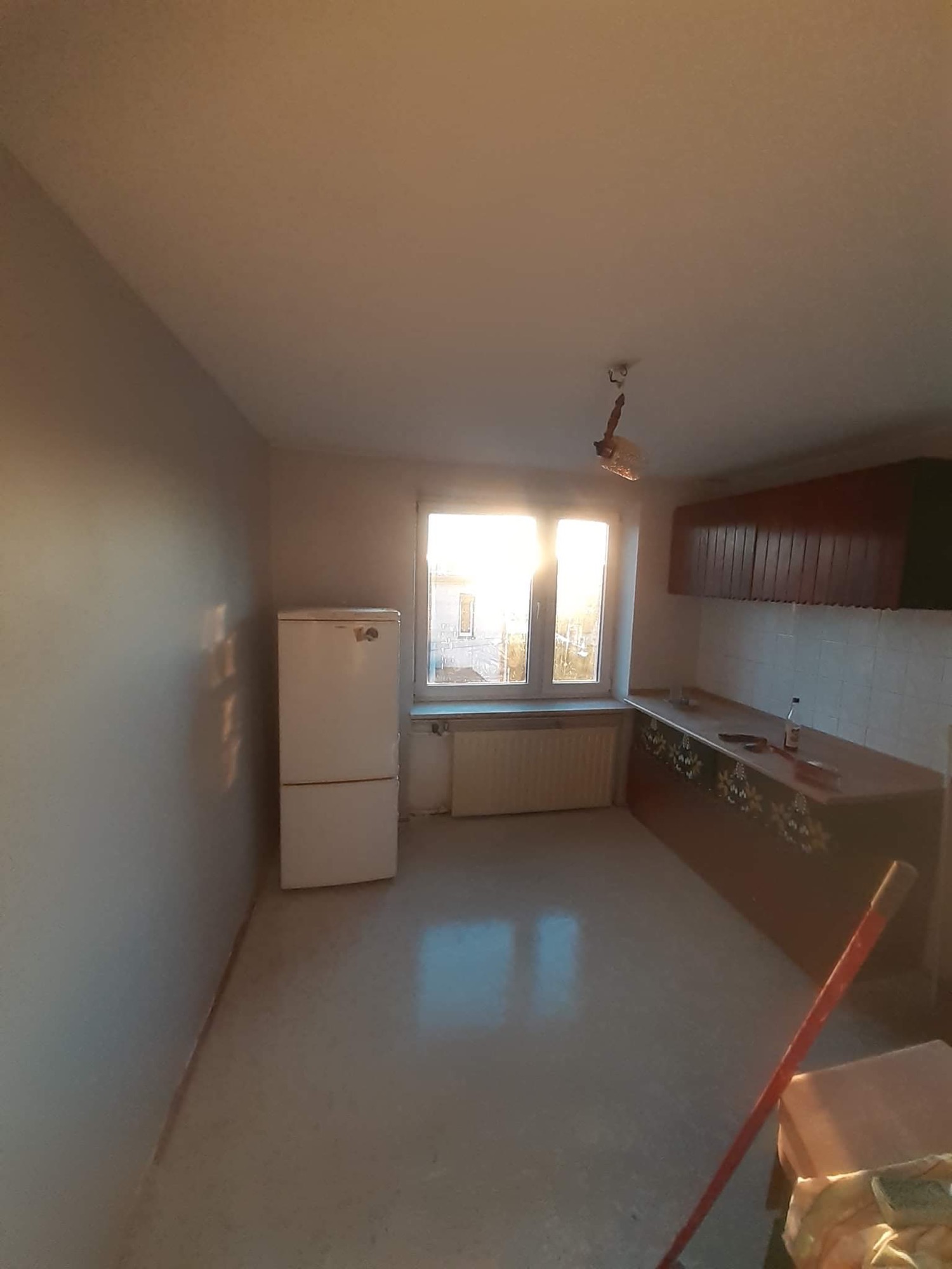 Wnętrze kuchni przed remontem: puste ściany, stara zabudowa kuchenna z dekoracyjnymi motywami kwiatowymi, biała lodówka i widok na okno z silnym światłem słonecznym.