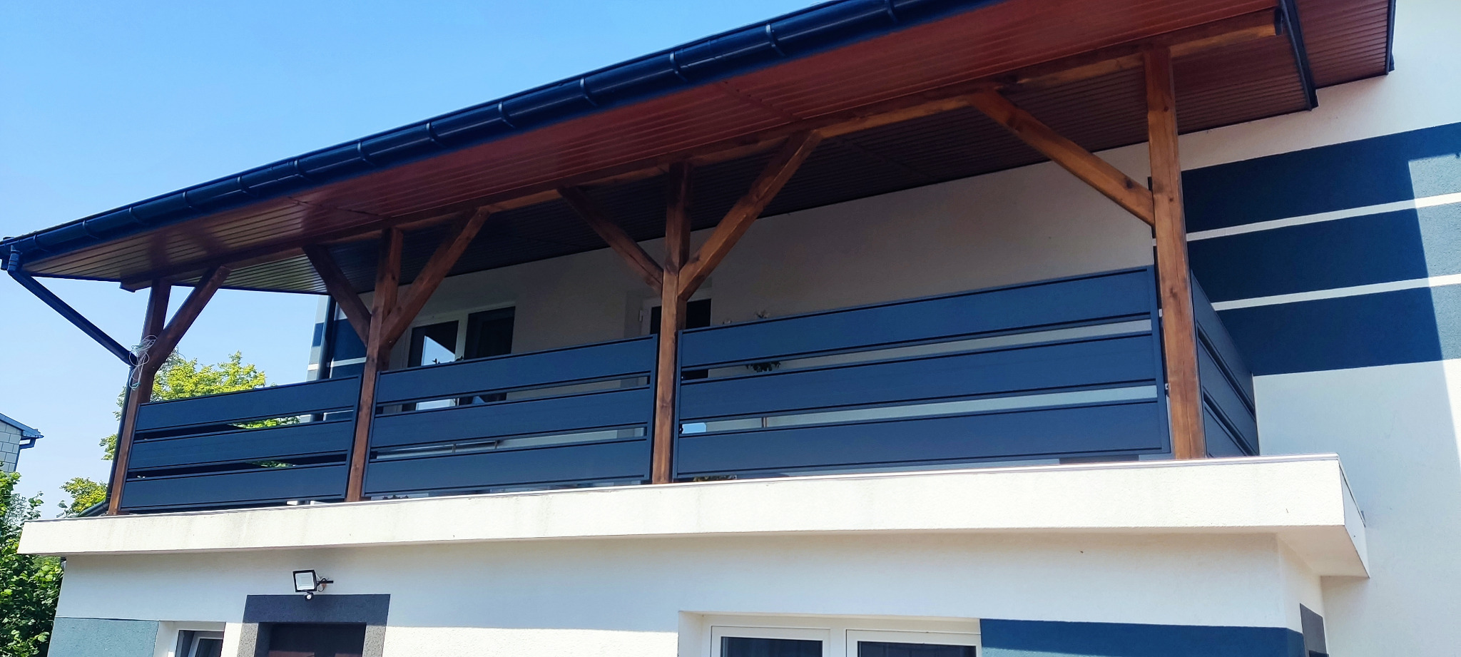 Nowoczesny balkon z poziomymi, grafitowymi panelami i drewnianą konstrukcją zadaszenia, widoczny z dołu, na tle błękitnego nieba.