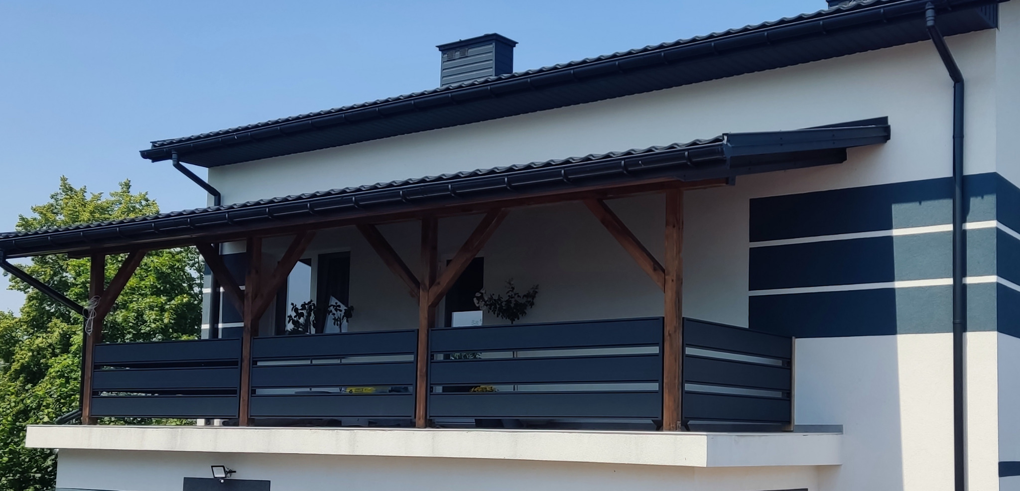 Nowoczesna balustrada balkonowa z poziomych, szarych paneli, wsparta na drewnianych słupach, z widokiem na zieleń i fragment elewacji budynku z ciemnymi pasami.