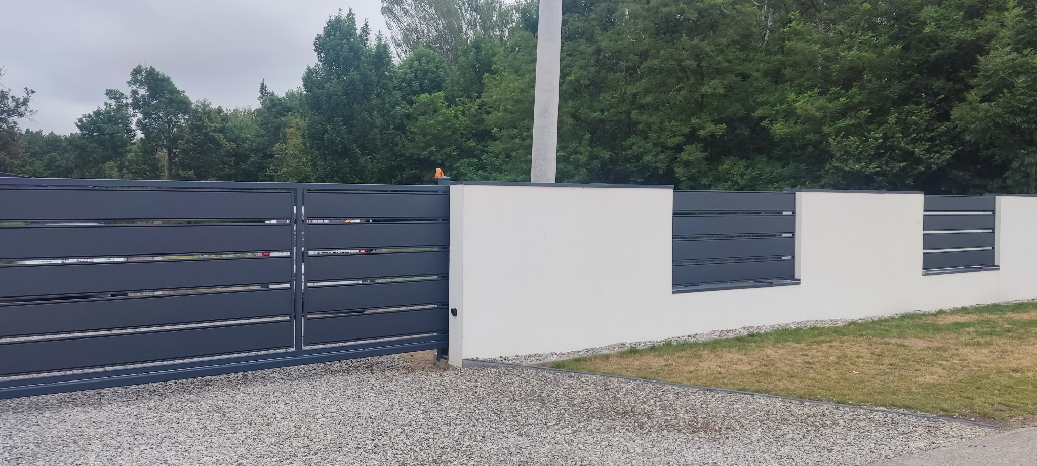 Nowoczesna posesja z przesuwną bramą wjazdową z poziomych profili w kolorze grafitowym, połączona z białym, murowanym ogrodzeniem z grafitowymi wstawkami, na tle zieleni drzew.