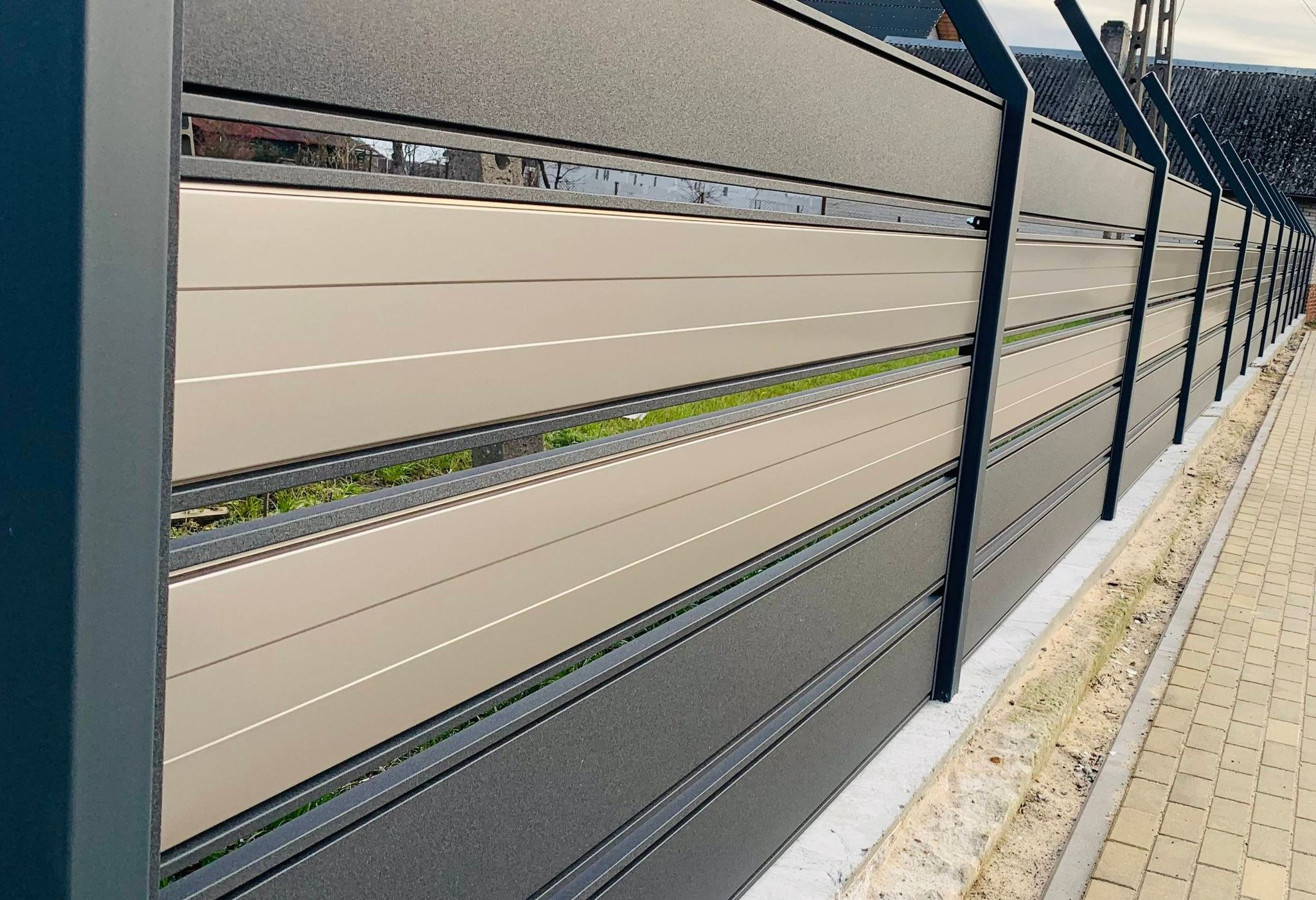 Nowoczesne ogrodzenie z paneli w odcieniach szarości i beżu, z poziomymi przerwami, widoczne z boku, z fragmentem chodnika z kostki brukowej i trawnika w tle.