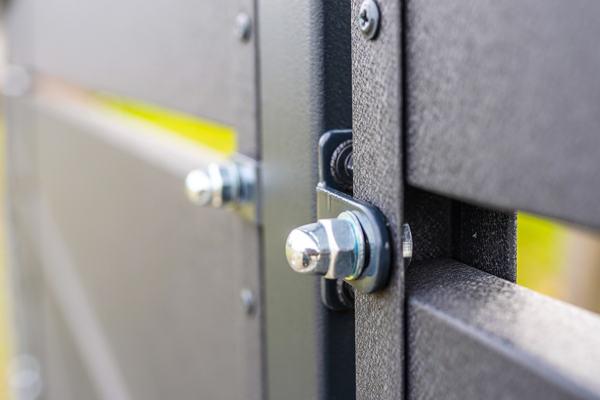 Szczegółowe zbliżenie na mocowanie śrubowe w metalowej konstrukcji, prawdopodobnie fragmentu bramy lub ogrodzenia; widoczne śruby, nakrętki i podkładki.