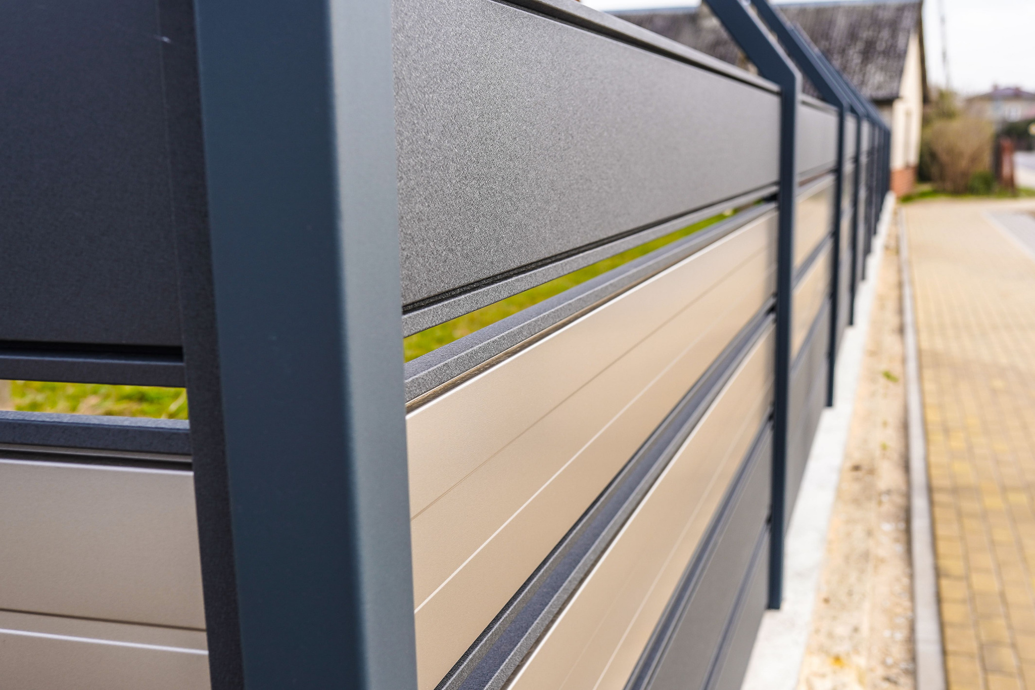 Nowoczesne ogrodzenie panelowe z poziomymi, beżowymi lamelami i grafitowymi słupkami, widok z bliska na tle kostki brukowej.