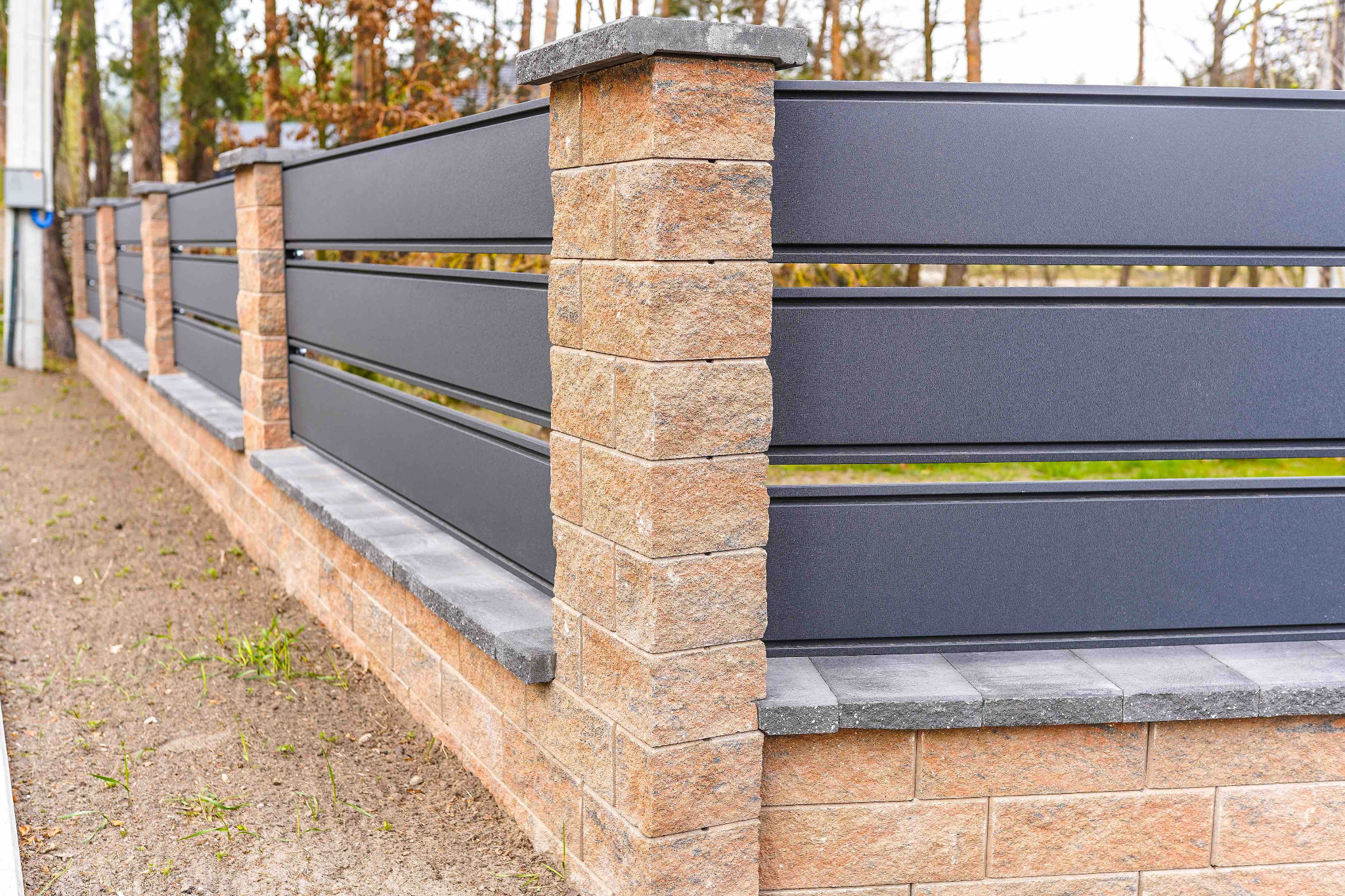 Nowoczesne ogrodzenie z szarych paneli poziomych i słupków z cegły klinkierowej z szarymi daszkami, widok z bliska na fragment ogrodzenia.