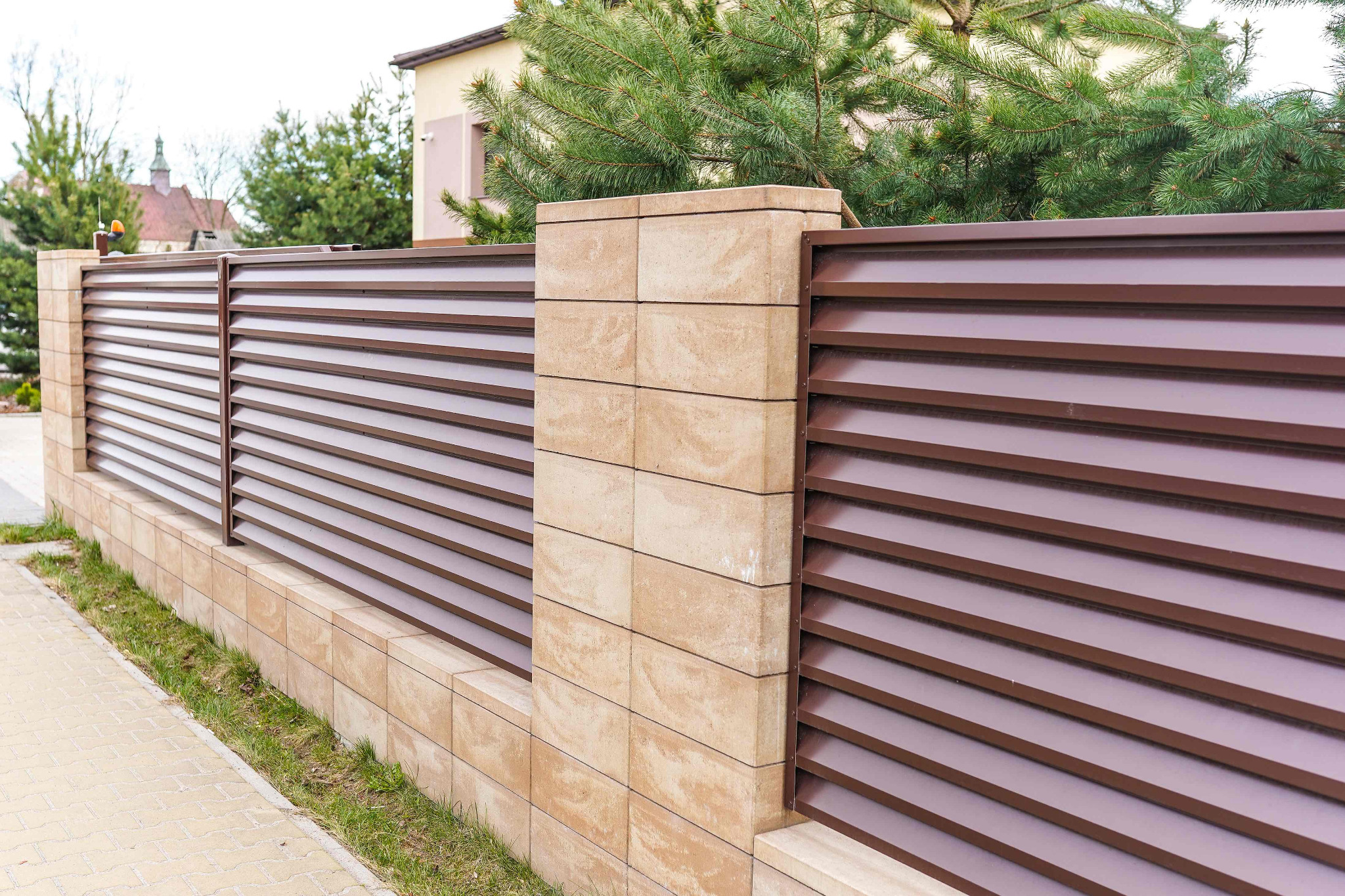 Nowoczesne ogrodzenie z poziomych, brązowych paneli zamontowanych na niskim murku z beżowych, prostokątnych bloczków betonowych, widok z perspektywy chodnika.
