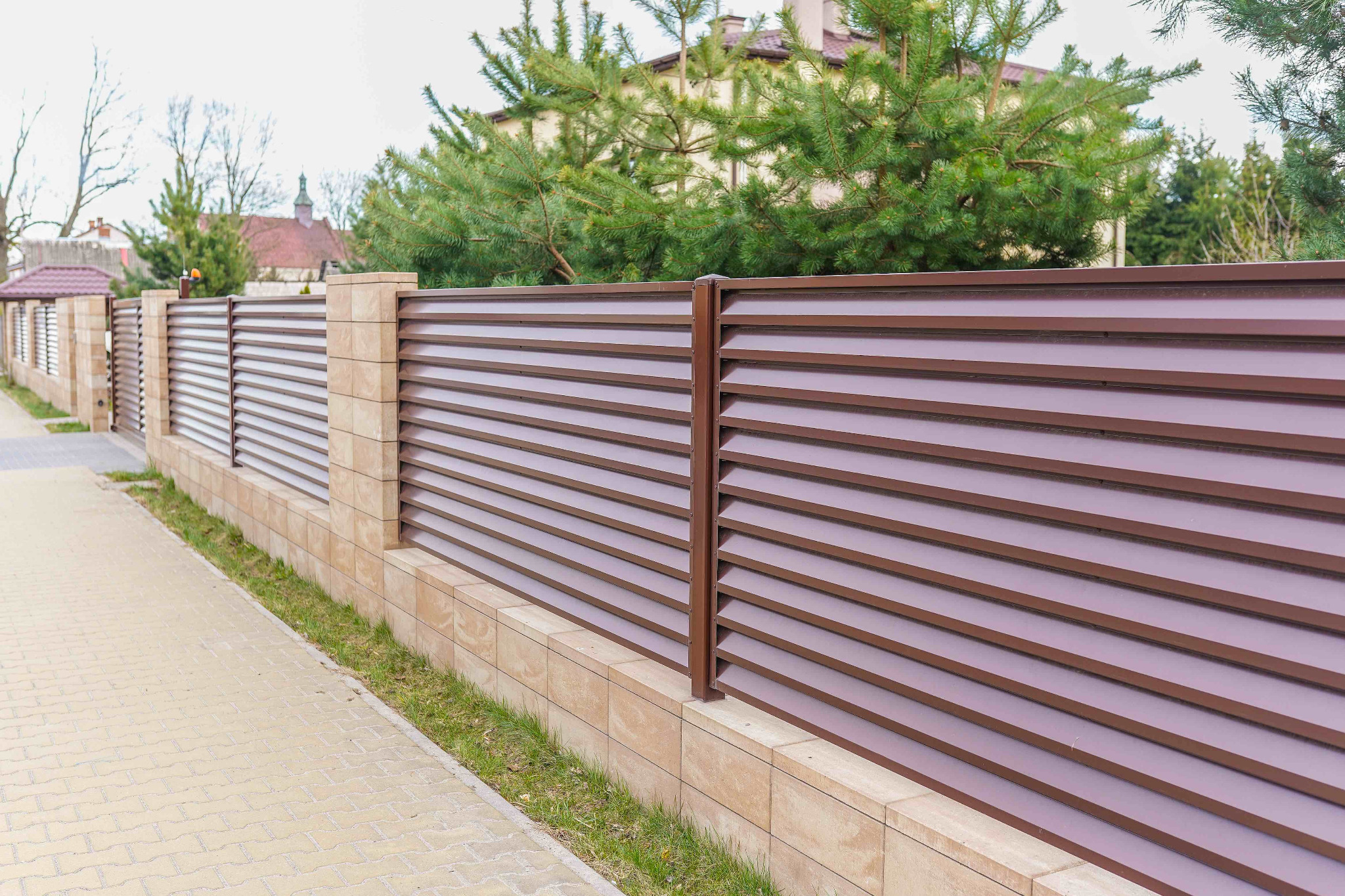 Brązowe ogrodzenie metalowe z poziomymi panelami i słupkami z piaskowca, widok z boku na tle zieleni i kostki brukowej.
