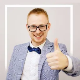 Uśmiechnięty mężczyzna w okularach i niebieskiej muszce, w jasnej marynarce w kratę, pokazuje kciuk w górę.