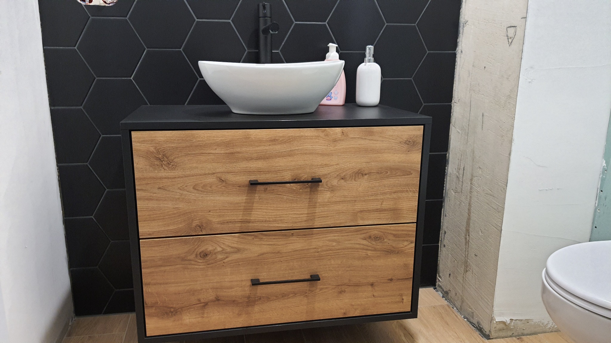 Nowoczesna umywalka nablatowa w kształcie misy na szafce z drewnopodobnymi frontami, czarna armatura i heksagonalne płytki w tle, widoczne niedokończone prace wykończeniowe na ścianie.