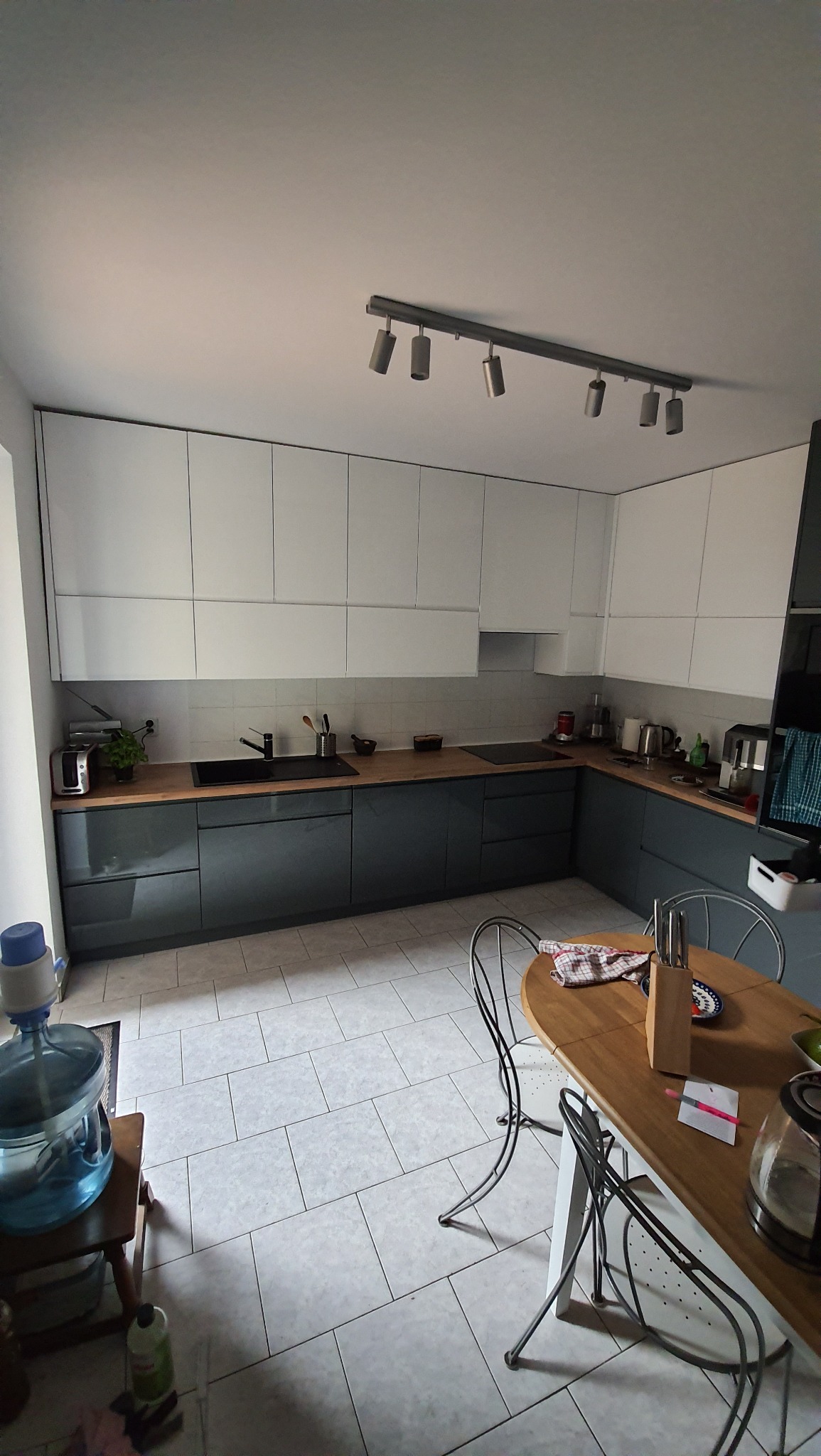 Nowoczesna kuchnia z białymi górnymi szafkami i grafitowymi dolnymi, drewnianym blatem, widoczne oświetlenie sufitowe punktowe i stół z krzesłami w tle.